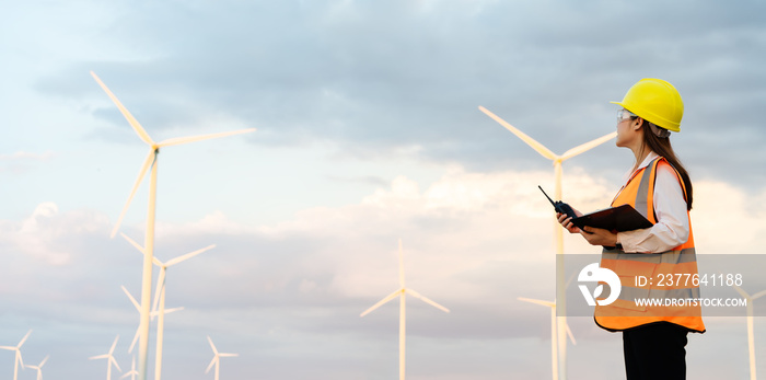 female engineer using walkie talkie and reading clipboard to checking system against wind turbine farm