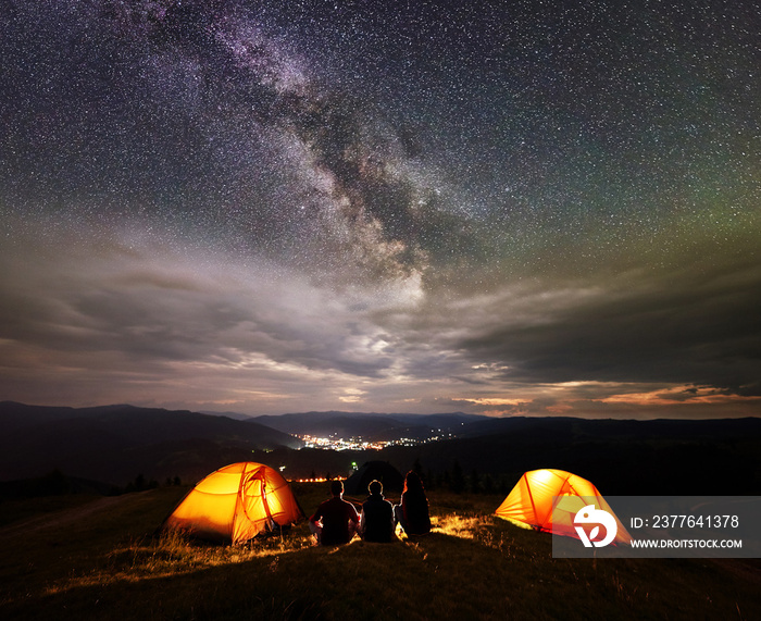 Rear view silhouette three tourists having a rest together beside camp, campfire and two orange tents at night on the top of mountain under wonderfull starry sky with many stars on background