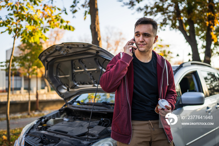 One young worried caucasian man standing by his vehicle with open hood and broken failed engine holding a phone calling towing service for help on the road Roadside assistance concept in autumn day