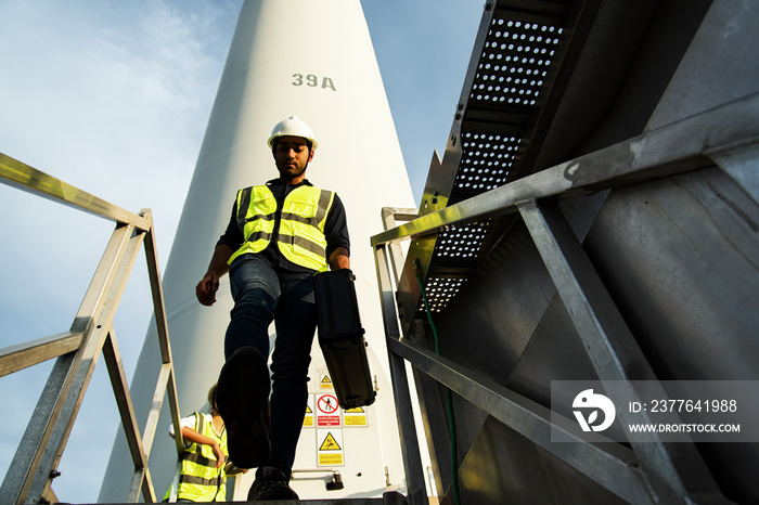 worker on a site. Young maintenance engineer team working with step down wind turbine farm on  sky background.