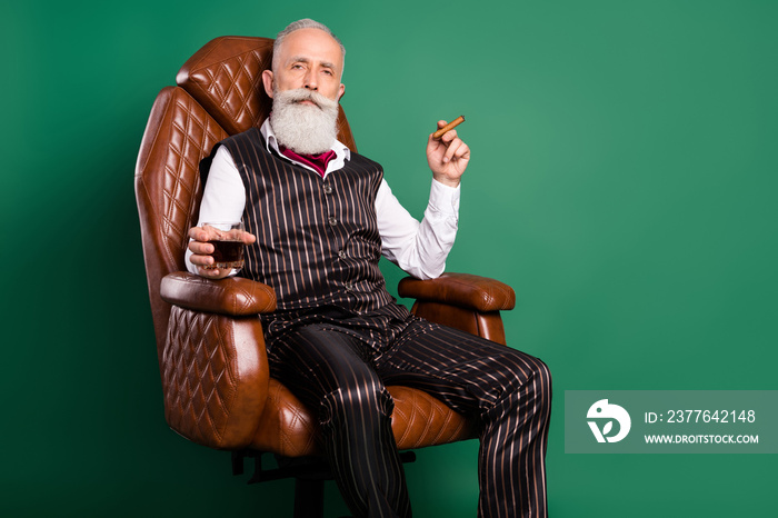 Photo of old noble aristocrat sit armchair drink whiskey enjoy cigar wear striped suit on green color background