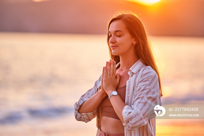 Portrait of calm serene satisfied woman with closed eyes and praying hands enjoys beautiful moment life on the seashore at sunset time