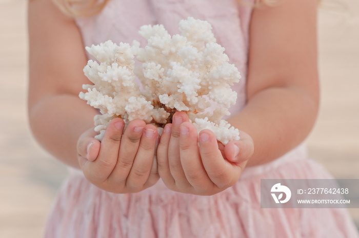 A little cute blonde girl in a gentle pink dress holds a coral in her hands by the sea. A beautiful baby girl plays with sea treasures. Decoration of celebrations, photo zones in elegant marine style