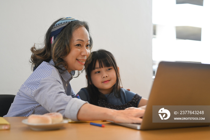 Happy mature grandma and little grandchild using laptop computer at home together. Multi generational, family and love concept