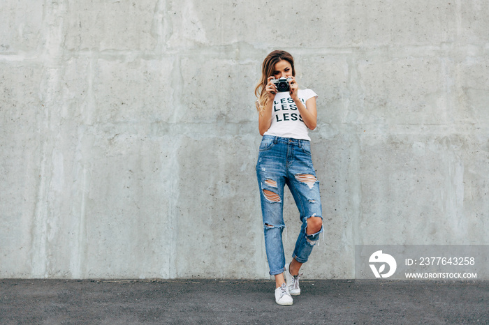 Stylish woman photographer in white T-shirt with word  less  with retro camera on the gray wall background. Image with copy space