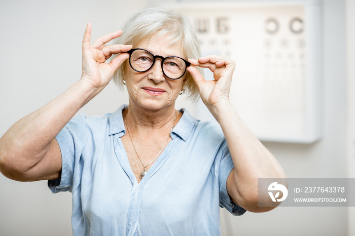 Portrait of a happy senior woman wearing eyeglasses in front of eye chart in ophthalmology office. Concept of checking eyesight and selecting glasses in older age