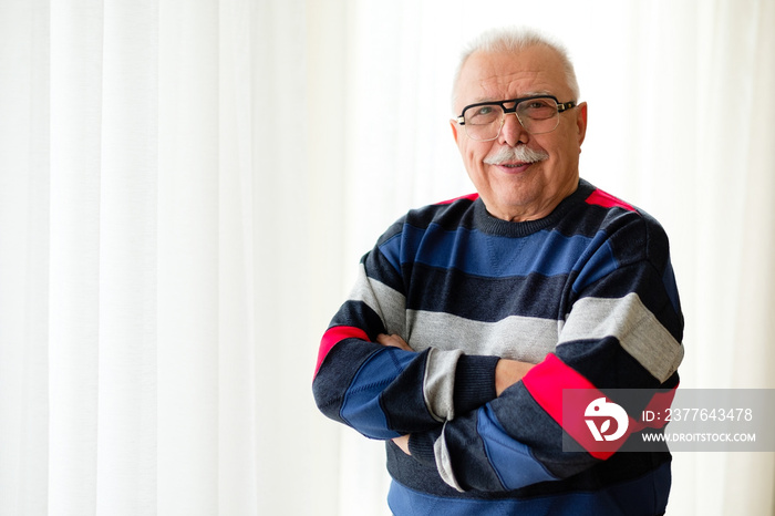 Portrait of smiling and confident with cross arms senior man 70-75 years old with eyeglasses stands near light window. Text space for ad