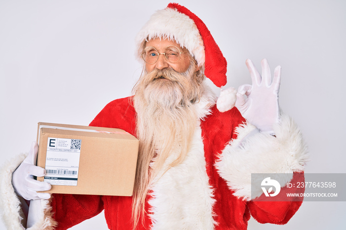 Old senior man with grey hair and long beard wearing santa claus costume holding delivery box doing ok sign with fingers, smiling friendly gesturing excellent symbol