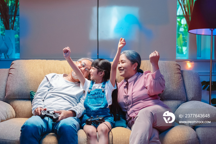 Asian happy family play time. little girl with grandparents together playing video games use joystick control enjoy laughing fun in living room home at night. Happy child happiness lifestyle concept.