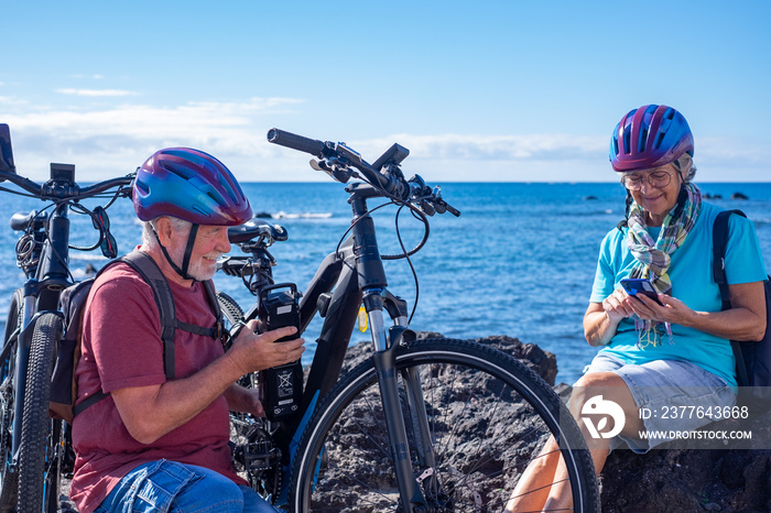 Caucasian carefree senior couple at the beach with electric bicycles in a sunny day. Man checks the battery while woman uses phone