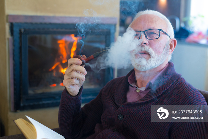 Senior man smoking pipe and reading book in front of the fireplace in winter