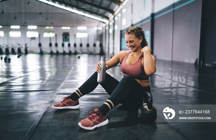 Cheerful fit girl dressed in tracksuit holding water bottle in hand and smiling during training day in gym, happy Caucaisan woman with protein cocktail resting in sportive studio - healthy lifestyle