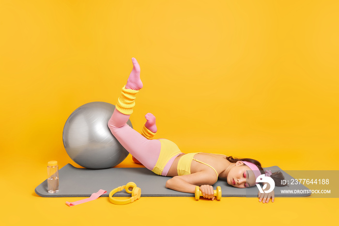 Exhausted sportswoman takes break after workout poses on fitness man with fitball headphones dumbbells bottle of water around isolated over yellow background. Healthy lifestyle and sport concept