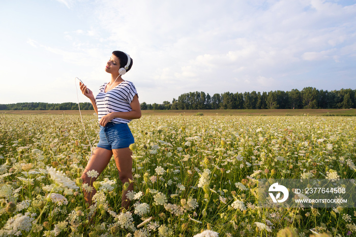 Young smiling woman wearing headphones listening music from her smartphone on the spring meadow full of white flowers