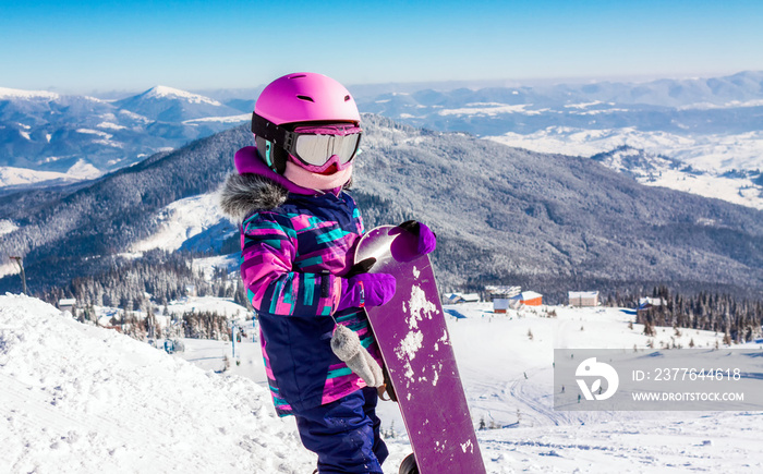 Little girl snowboarding with equipment helmet and goggles outwear holding snowboard resting on top of ski slope in sunlight