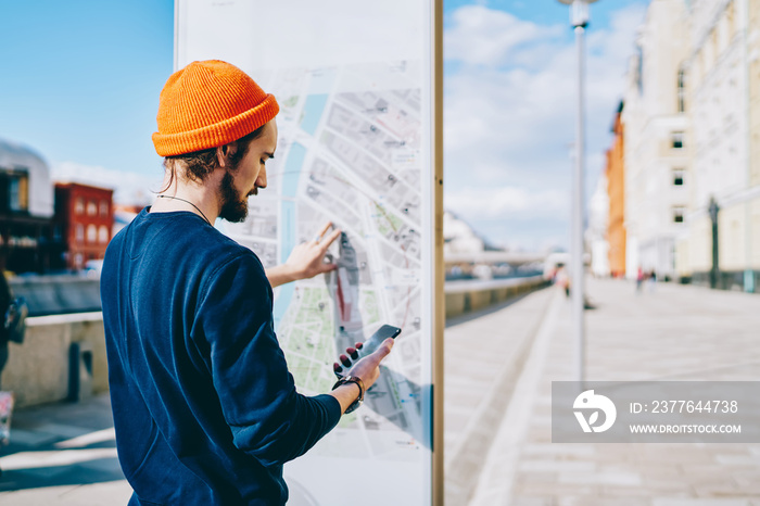 Hipster guy checking schedule of public transport on modern electronic board with information, back view of male traveler searching location on mobile phone application and on map board on street.