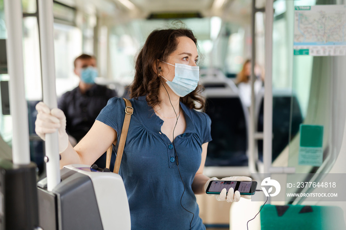 Portrait of female passenger in disposable face mask and latex gloves using mobile phone while traveling by city streetcar. New life reality during COVID 19 pandemic