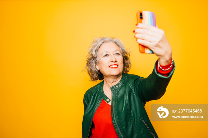 Senior woman taking a selfie with a smartphone with the rainbow flag on the case, over a yellow background