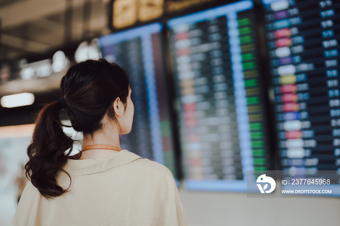 Young asian woman with passport and boarding pass as a hand in international airport looking at the flight information board, checking her flight