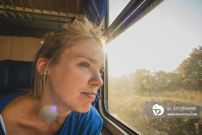 Caucasian woman using looking through a window of an older train with vintage seats and wooden walls. Woman commuting with an older retro train. Sun flare visible