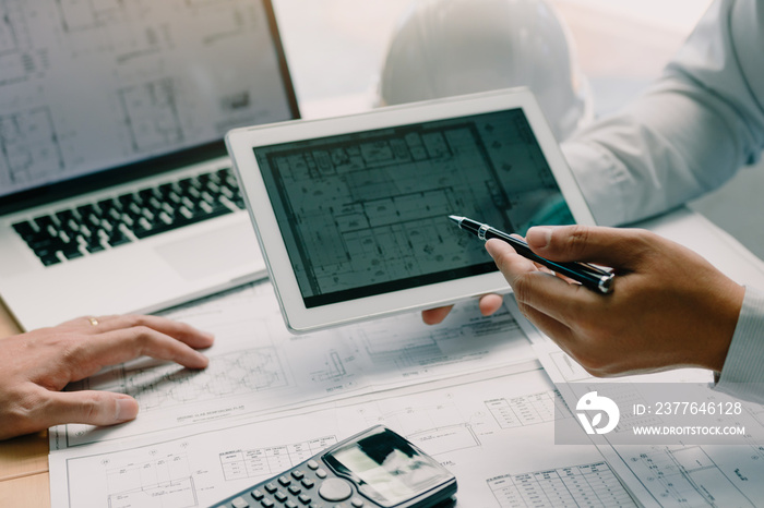 Two engineering working together and using digital tablet looking blueprint and analysis with architectural plan on desk.