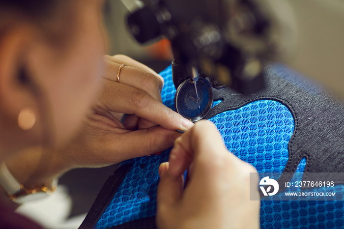 Shoe factory worker making black and blue sneakers using industrial sewing machine, hands holding shoe detail in close up. Footwear manufacturing industry concept