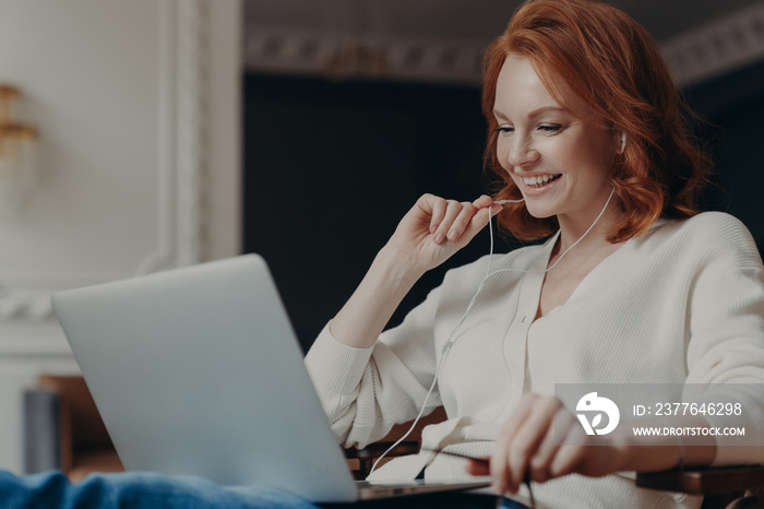 Positive redhead woman studies online course, talks with friend in application, uses earphones, enjoys video conference from favorite blogger, browses internet. Distant communication and work