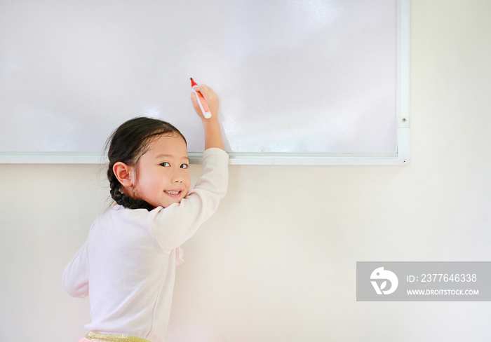 Portrait of happy little Asian child girl or Schoolgirl writing something on whiteboard with a marker and looking at camera in the classroom. White board with copy space for text.