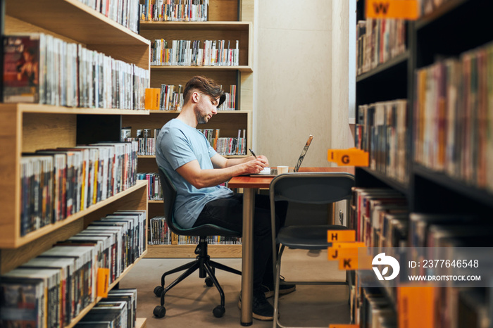 Student learning in library. Young man preparing for test on laptop. Man listening to online course. Focused student studying for college exams