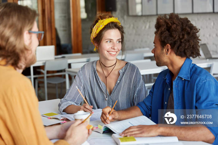 Friendly atmosphere and good relationships. Multiracial students doing their home assignment sitting in classroom. Young pretty student female trying to help her groupmate to catch with studying