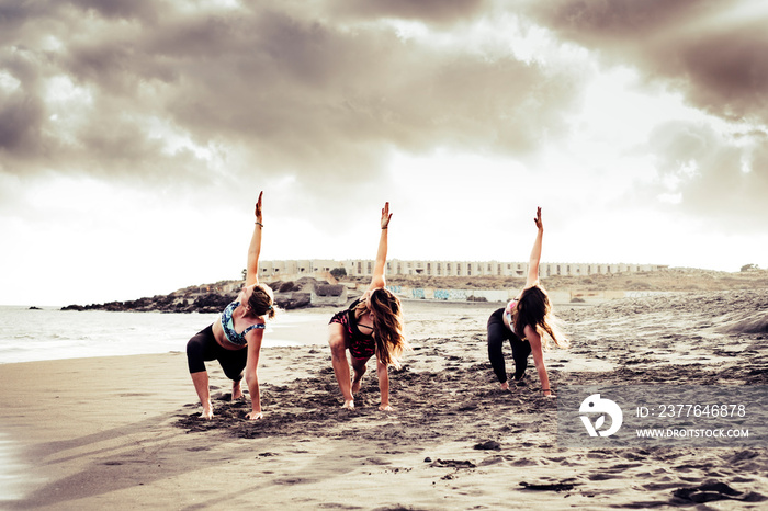 Pilates balance position three young active women at the beach doing sport fitness activity together under a dramatic cloudy sky - scenic image of people doing sport