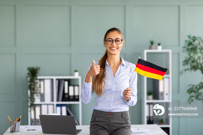 Happy female tutor with flag of Germany showing thumb up, standing near workplace in office interior