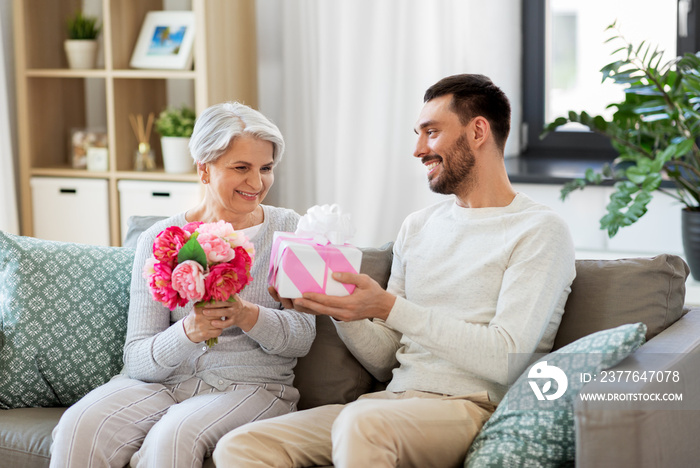 family, mother’s day and birthday concept - smiling adult son giving present and flowers to his senior mother at home