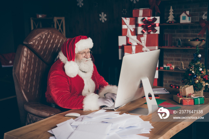 Profile side photo of concentrated jolly grey beard santa claus writing kids x-mas christmas wish list use computer wear red cap headwear sit table in house indoors newyear decoration