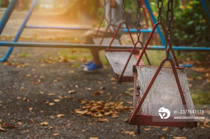 Blurred alone boy sitting on old wooden garden swings in abandoned playground with sunset lighting background, Selective focus at the front wood seat.