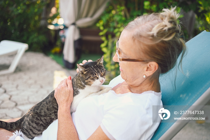 Happy smiling senior elderly woman in glasses relaxing in summer garden outdoors hugging domestic tabby cat. Retired old people and animals pets concept