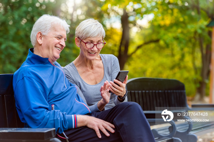 Smiling senior active couple sitting on the bench looking at smartphone