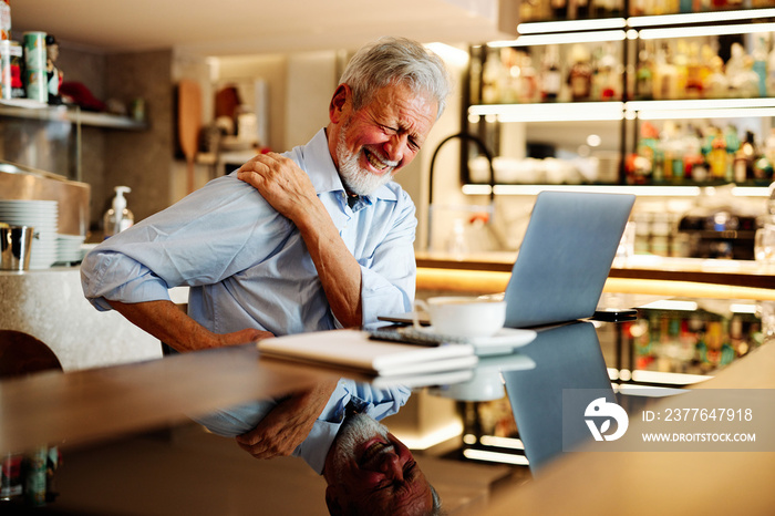 A senior man sits in a coffee shop and has back pain.