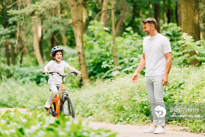 full length view of son riding bicycle and father standing near boy and looking at kid