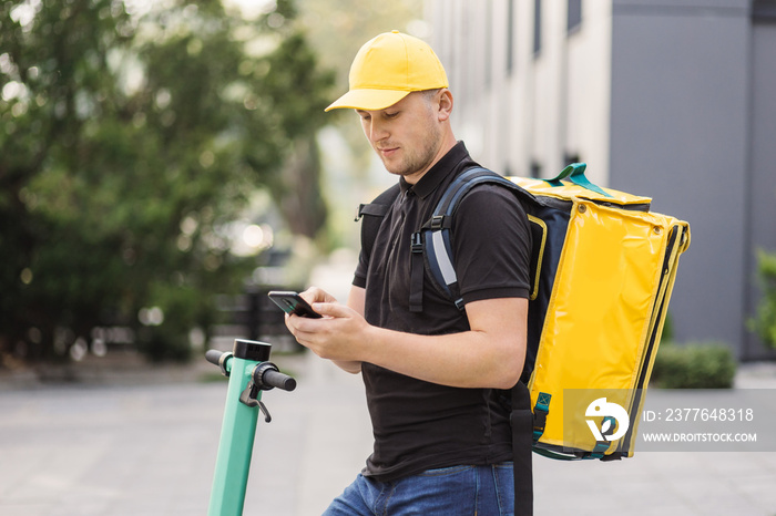 Courier man is looking in mobile phone to deliver address of food from a restaurant. Food delivery guy navigates in phone in city with electric scooter. Eco friendly transportation.