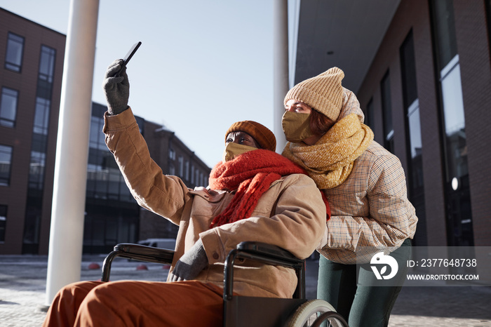 Side view portrait of African American man using wheelchair and wearing mask while taking selfie photo with young woman assisting in urban city, copy space