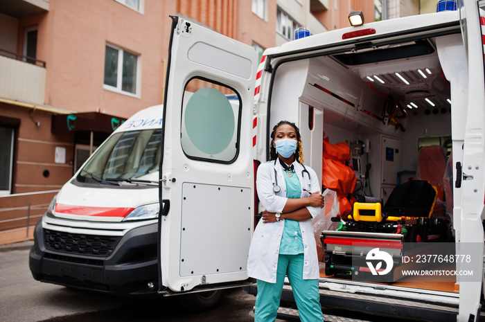 African american female paramedic in face protective medical mask standing in front of ambulance car.