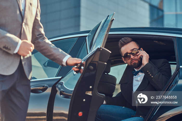 Attractive groomed man in sunglasses is talking by smartphone and sitting in the car while his assistant is opening door for him.