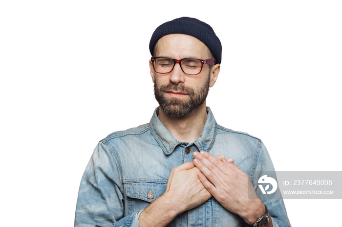 Pleased bearded man with closed eyes, keeps hands on chest, dressed in fashionable clothes, isolated over pink studio background. Middle aged male with shut eyes, expresses his love and sympathy