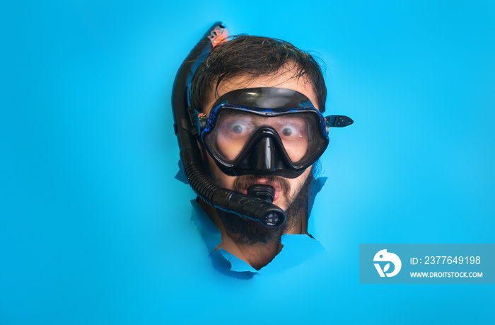 Happy funny man in a swimming mask, with snorkel in his mouth through torn paper blue background.