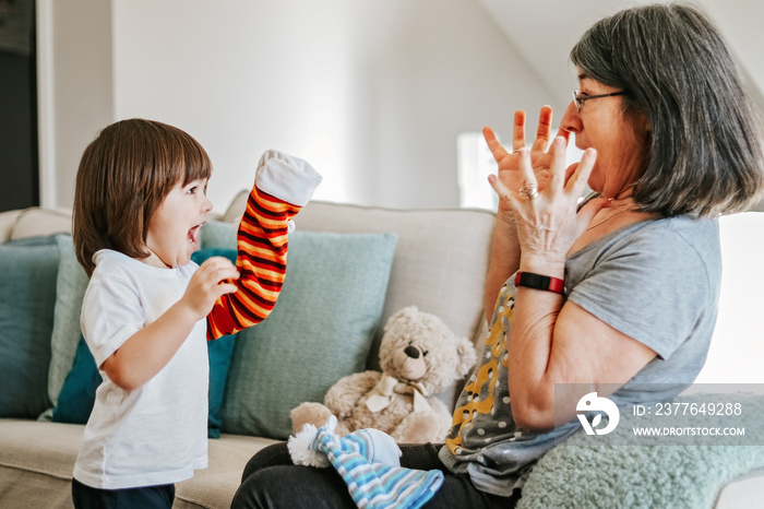 Cute little cheerful child playing with his older granmother with bright hand toy at home. Focus on boy. Family lifestyle. Active senior woman. Babysitting.