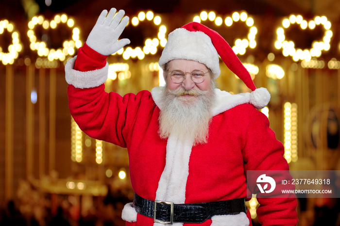 Santa Claus smiling and waving. Happy bearded Santa Claus raised hand in greeting gesture on New Year lights background. Santa Claus on blurred background.