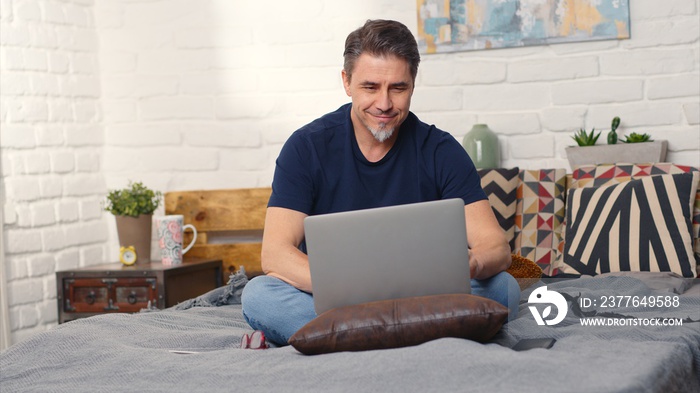 Stay at home and work in home office - Casual man sitting on bed working online from home with laptop computer.