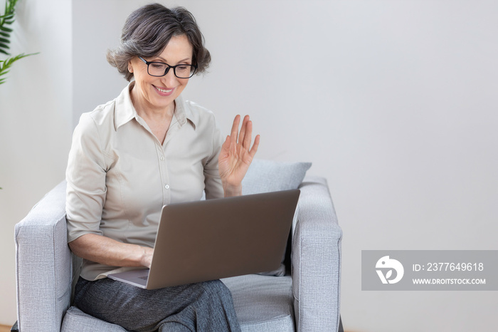 Beautiful caucasian aged business woman using laptop computer for online communication via the internet sitting on an armchair in the office or at home. Video call, remote work concept