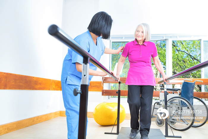 Asian nurse assisting aged senior woman training on the bars at rehab facility